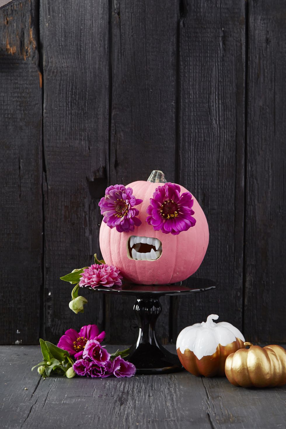 Pink pumpkin with flower eyes and playful vampire teeth, set against a backdrop of dark brown wooden flooring and walls